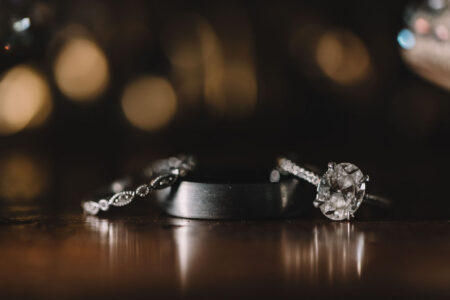Diamond engagement ring, dark wedding band, and delicate stone band on wood with soft, warm blurred lights in background.