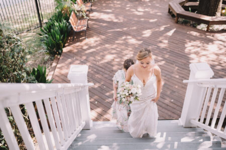 Bride in white dress climbs outdoor stairs with bouquet, followed by woman in floral dress; sunlight shines on wooden deck below.