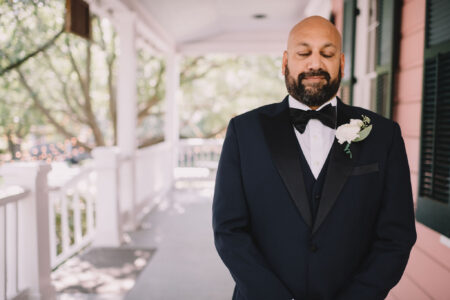 A man in a tuxedo with a white boutonniere stands calmly on a porch, eyes closed, surrounded by railings and trees.
