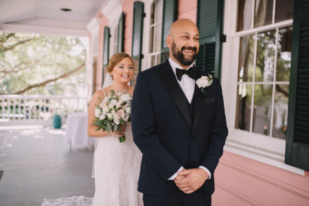 Bride in white dress with bouquet smiles behind groom on porch with pink walls and green shutters, ready for first look.