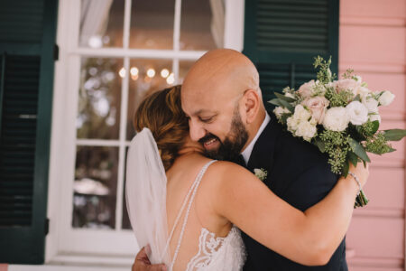 Bride in white dress hugs smiling man with bouquet by window with green shutters.