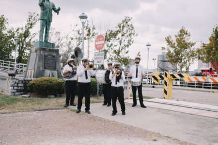 Six brass musicians in white shirts and black pants perform outdoors near a Do Not Enter sign, statue, and cloudy skies.