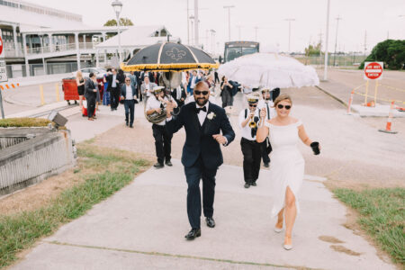 Smiling bride and groom with decorated umbrellas lead guests in a parade; groom in tux, bride in white, some guests play instruments.