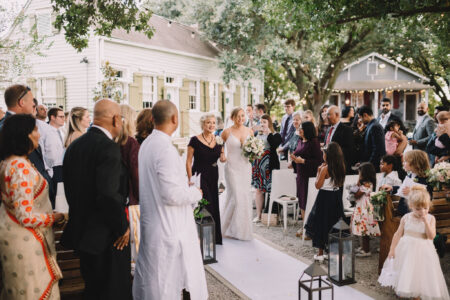 Bride in white holding hands with older woman walks outdoor aisle as formal guests watch, under trees and string lights.