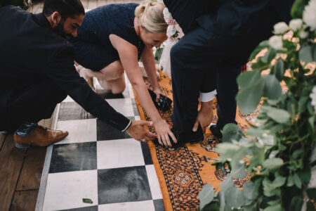 Three formally dressed people help adjust a man’s shoe on an orange rug beside a checkered floor and green foliage.
