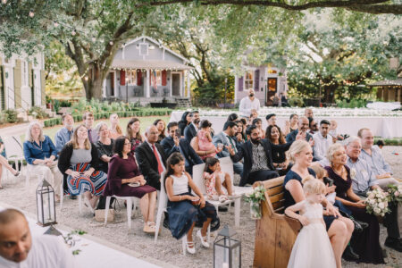 Guests smile and watch attentively at an outdoor wedding under string lights, with trees and houses in the background.