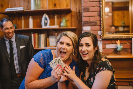 Two women smile and hold cash, with a suited man smiling behind them in a cozy, wood-paneled room with shelves and a mirror.