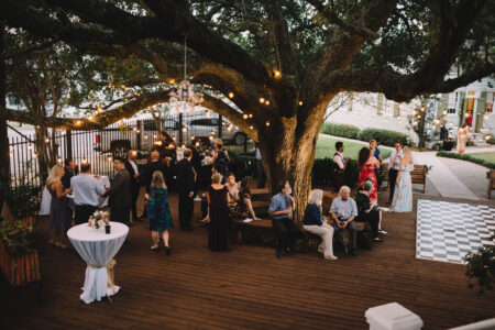 People gather under a tree with string lights on a deck for an outdoor event, near tables and a checkered dance floor.