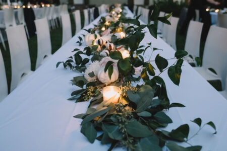 Elegant outdoor dinner setup with a white table, greenery, white flowers, and glowing candles in glass holders.