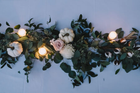 Greenery, white mini pumpkins, a pale pink rose, white flowers, and three candles on a light blue tablecloth.