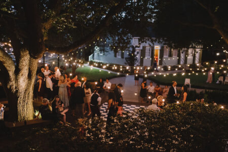 Guests celebrate outdoors at night under string lights, with a checkered dance floor, trees, and a house in the background.