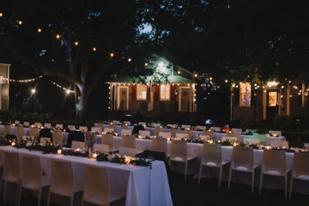 Outdoor banquet tables with white chairs, candles, and greenery under string lights; lit house in background at night.