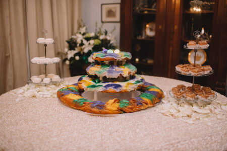 King Cake with purple, green, and yellow icing sits on a table with cookies, fudge squares, and scattered white petals.