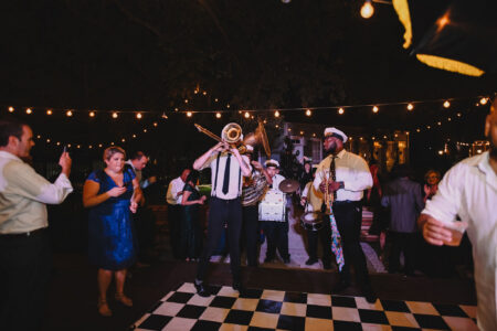 Outdoor night party with a live brass band, people dancing, and string lights over a black-and-white checkered dance floor.
