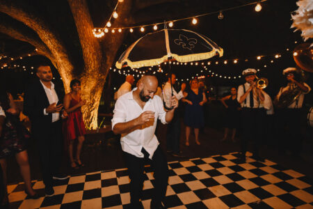 Man dancing with black and gold umbrella on checkered floor, surrounded by people and musicians at a festive outdoor night event.