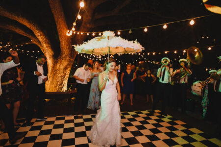 Bride in white dress dances with feathered umbrella on checkered floor, surrounded by guests, brass band, and string lights.