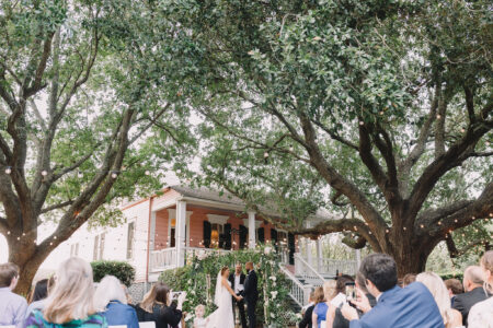 Couple exchanges vows at an outdoor wedding under large trees by a pink house, surrounded by guests.