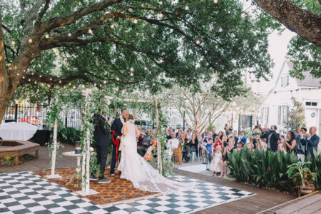 Bride and groom under a decorated outdoor arch, greenery and string lights, as guests watch the ceremony on a checkered floor.