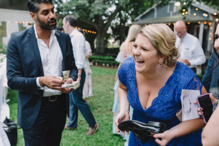 Woman in blue dress laughs holding a shoe as suited man counts money; others and string lights in outdoor setting.