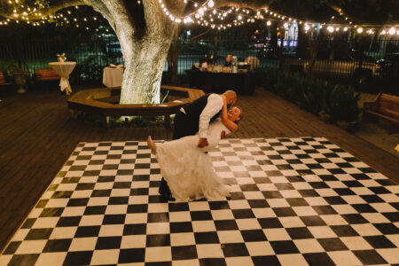 Bride and groom dance on a checkered floor under string lights, with a large tree and tables in the background.