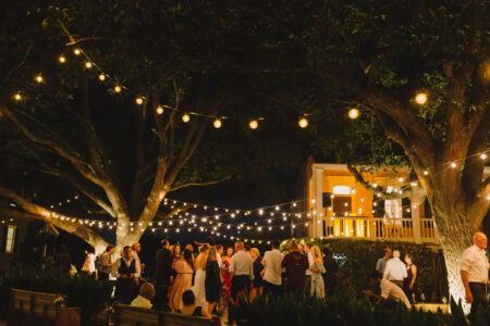 Outdoor night gathering under string lights, near large trees and a warmly lit house, festive and celebratory mood.