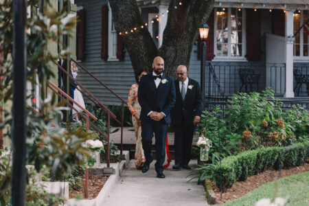 Man in tuxedo walks arm in arm with older man and woman on a garden path, string lights and house behind them.