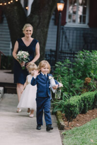 Boy in a blue suit waves on a garden path, holding a jar and stuffed animal; girl and woman with flowers follow.