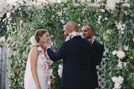 Bride and groom smile under a floral arch as the groom touches her face; officiant stands behind amid white flowers and greenery.
