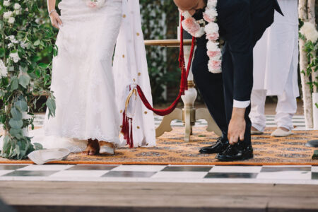 Bride and groom in traditional attire perform a wedding ritual on a rug, linked by red cloth, with greenery and barefoot bride.