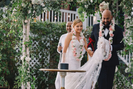 Bride and groom with flower garlands smile as they walk under a floral arch at an outdoor wedding, holding the wedding gown.