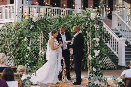 Bride and groom exchange vows under a floral arch at an outdoor wedding, with guests seated and string lights overhead.