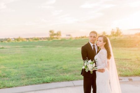 Bride in white lace dress and veil with groom in black suit, bouquet in hand, outdoors at sunset on a sunny Louisiana day.