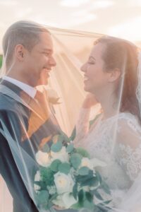Bride and groom smiling under a sheer veil, bride holding white roses, in romantic New Orleans sunlight.