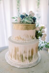 Elegant two-tier white wedding cake with gold brushstrokes, white flowers, greenery, and pearl accents on a round board.