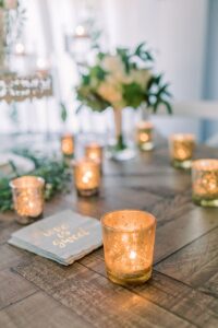 Gold votive candles and “love is sweet” napkins on wood table; blurred floral decor in background at a wedding reception.