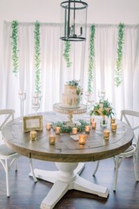 Wood table with white base holds two-tiered cake, gold candles, flowers, and sign; greenery and lantern light in background.