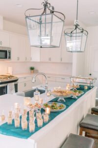 White kitchen island set for a Gulf South wedding, with candles, appetizers, snacks on a teal runner, and elegant lantern lights overhead.