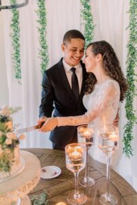 Bride and groom smiling, embracing as they cut their Gulf South wedding cake amid white drapery, greenery, and floating candles.