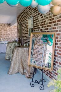 Framed welcome sign with white script on an easel beside gold tables, teal and gold balloons, at a brick-walled Gulf South wedding.