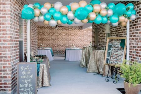 Stylish Louisiana event space with teal, gold, and white balloons, gold tablecloths, brick walls, and elegant wedding decor.