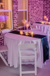 Romantic wedding table with white cloth, dark runner, candles, white chairs, brick walls, and purple-lit windows.