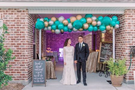 Create a dramatic entrance to your cocktail hour on the patio with a balloon arch, mirror welcome signage and sparkly sequin linen. Bride and groom hold hands under a colorful balloon arch at a festive, warmly lit New Orleans wedding entrance.
