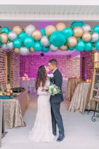 Bride and groom kiss under teal, gold, and silver balloons at a sparkling Gulf South wedding with gold tables and purple lighting.