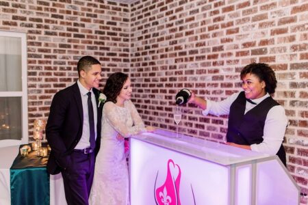 A newlywed couple smiles as a bartender pours drinks at a stylish, modern bar with a brick wall—perfect Louisiana wedding inspo.