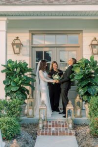 Bride and groom laugh, holding hands at their Louisiana outdoor wedding, officiant between them, lanterns and plants at entrance.