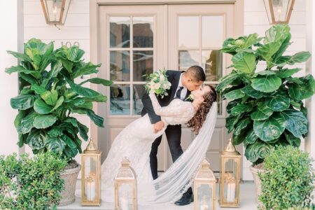 Groom in black suit kisses bride in white gown outside doors, with green plants and gold lanterns at New Orleans wedding.