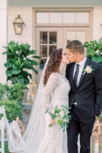 Bride in lace gown and veil kisses groom in black suit, holding white bouquet outside with potted plants and lanterns.