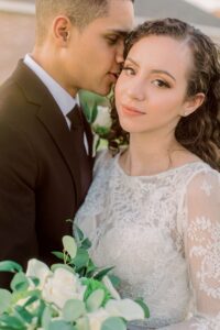 Groom in black suit kisses bride’s temple; she holds a white bouquet at their elegant New Orleans wedding.