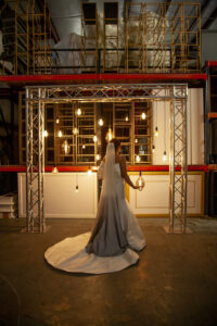 Bride in white dress and veil stands under hanging lights in an industrial setting with metal shelves and concrete floor.
