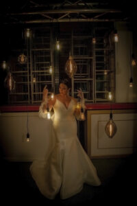 Bride in white gown touches glowing vintage bulb in a dark room, capturing a magical New Orleans wedding moment.